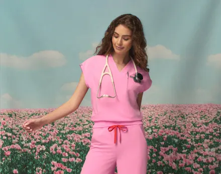 Healthcare professional wearing Sunrise Pink scrubs standing in a blooming flower field, highlighting the new seasonal color from the FIGS Spring Drop.