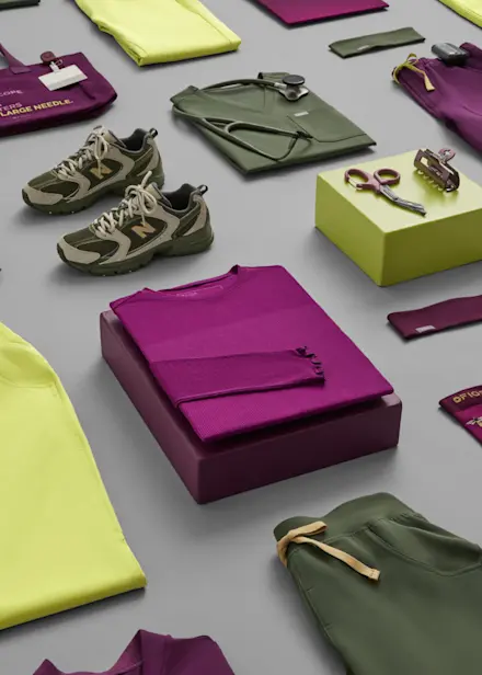 Flat lay of FIGS scrubs and healthcare accessories in Celery, Deep Purple, and olive green, including folded uniforms, tote bag, stethoscope, badge, and shoes.
