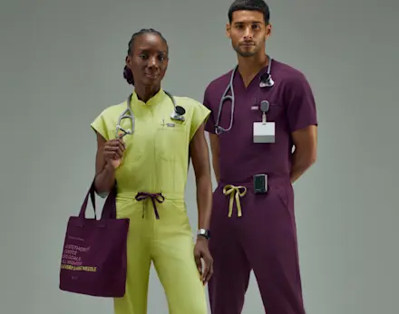 Two healthcare professionals wearing FIGS FlexKnit scrubs in Celery and Deep Purple stand side by side with medical accessories, showcasing the new color collection.