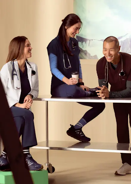 Four healthcare professionals wearing FIGS scrubs gather around a table, smiling and talking during a break in a bright, modern workspace.
