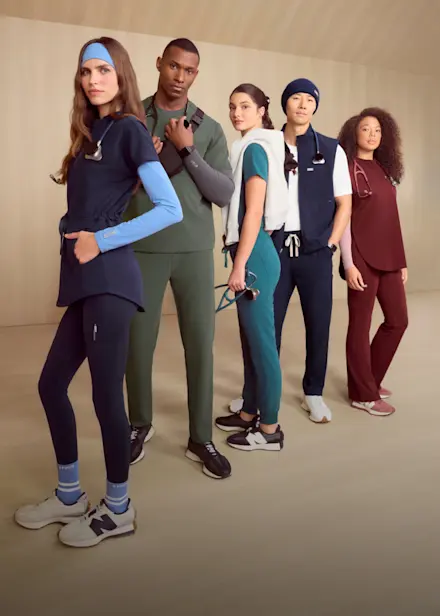 Five healthcare professionals wearing colorful FIGS scrubs stand together in a minimalist studio, showcasing new 2026 scrub styles and accessories.