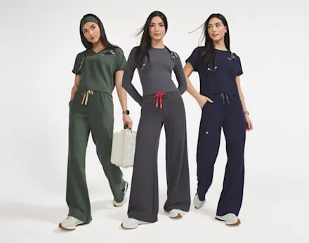 Three women wearing FIGS cropped scrub tops and wide-leg scrub pants in Moss, Charcoal and Navy, showcasing the new silhouette collection.