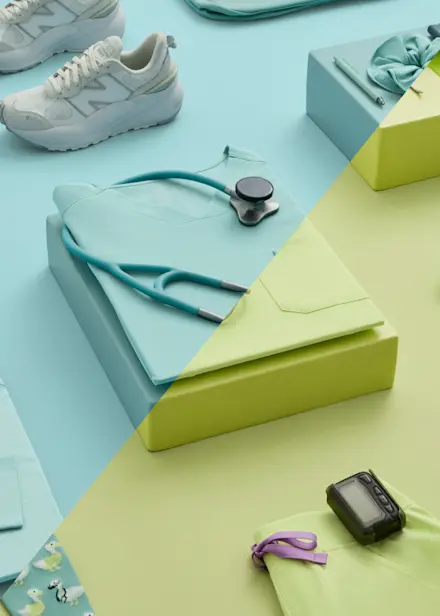 Diagonal flat lay of Seaglass and Sea Mist FIGS scrubs with a stethoscope displayed on coordinating blue and green backgrounds.