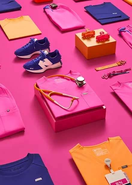 Flat lay featuring FIGS Nurses Week collection with Deep Royal Blue New Balance sneakers, colorful scrub apparel, healthcare-inspired accessories, and campaign merchandise.